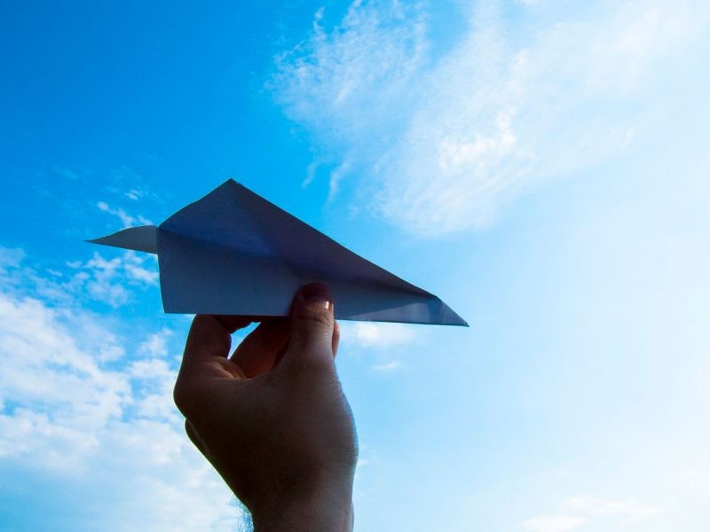 Mano que sujeta un avión de papel y de fondo el cielo despejado con alguna pequeña nuve blanca